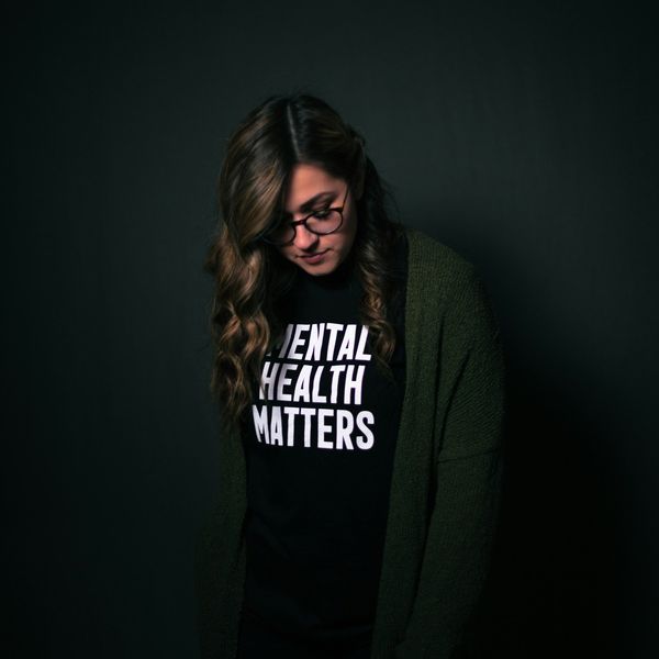 young woman standing alone in a dark room with a shirt that reads "Mental health matters".