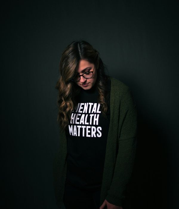 young woman standing alone in a dark room with a shirt that reads "Mental health matters".