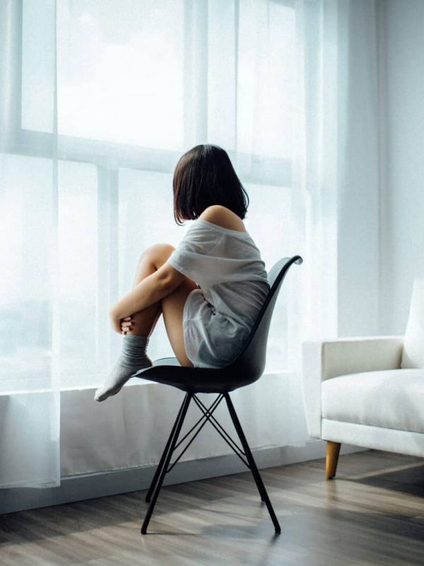 young girl sits in a chair alone by a window as she struggles with mental health issues