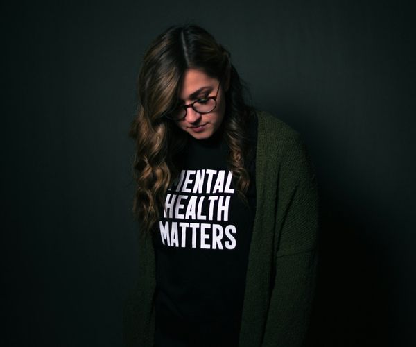 young woman standing alone in a dark room with a shirt that reads "Mental health matters".